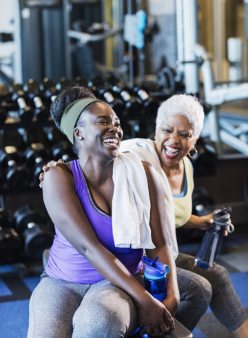 A senior African-American woman in her 60s exercising at the gym with her adult daughter, a young woman in her 20s. They are taking a break from lifting dumbbells, conversing and drinking from water bottles.