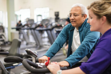 Older gentleman riding a stationary bike with a female trainer assisting